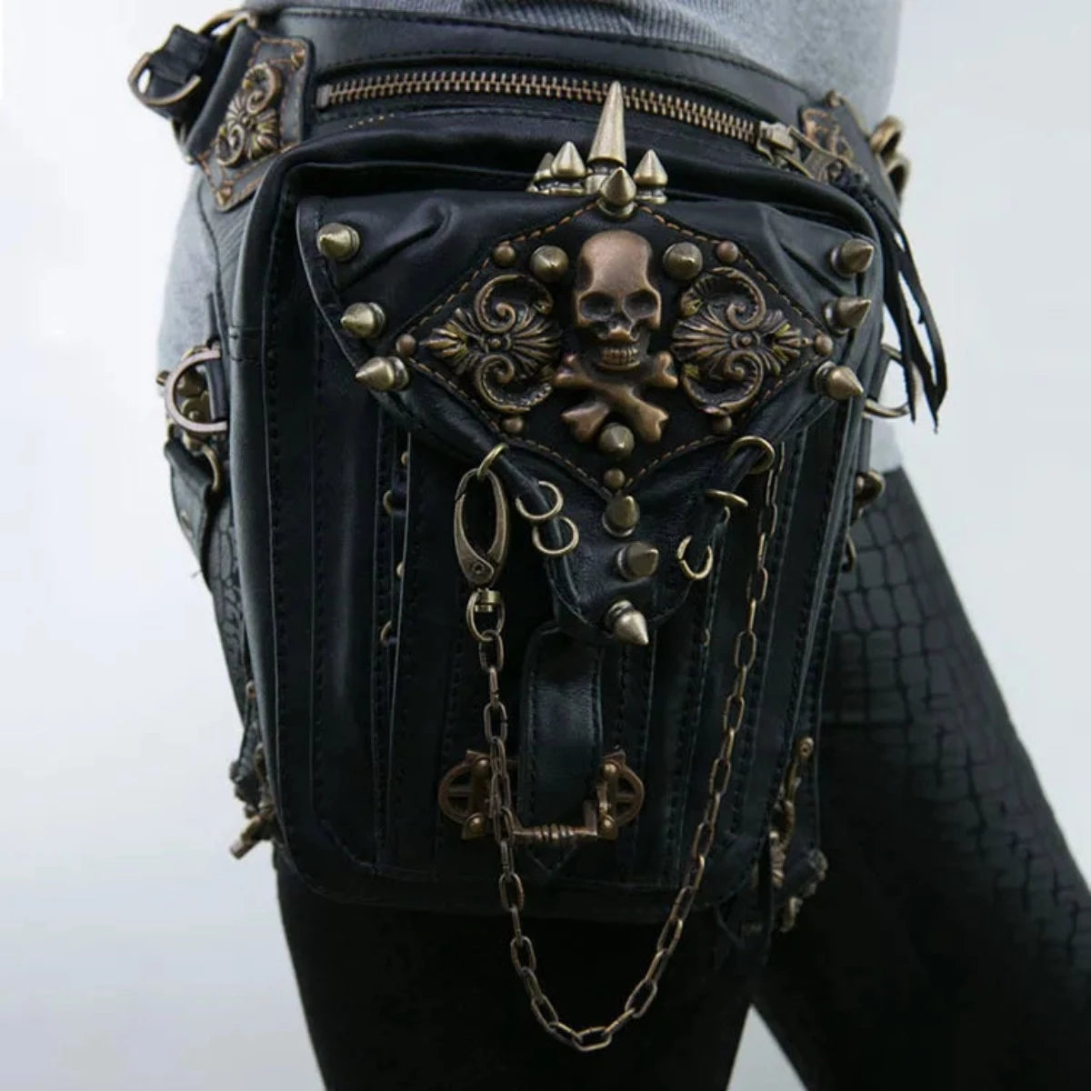 A Women's Gothic Waist/Hip/Body Fanny Bag in black leather, featuring ornate bronze skull and crossbones detail, chains, and metal studs. Worn on the side of a person sporting textured black leggings, this Gothic-inspired design offers spacious compartments for all your essentials.