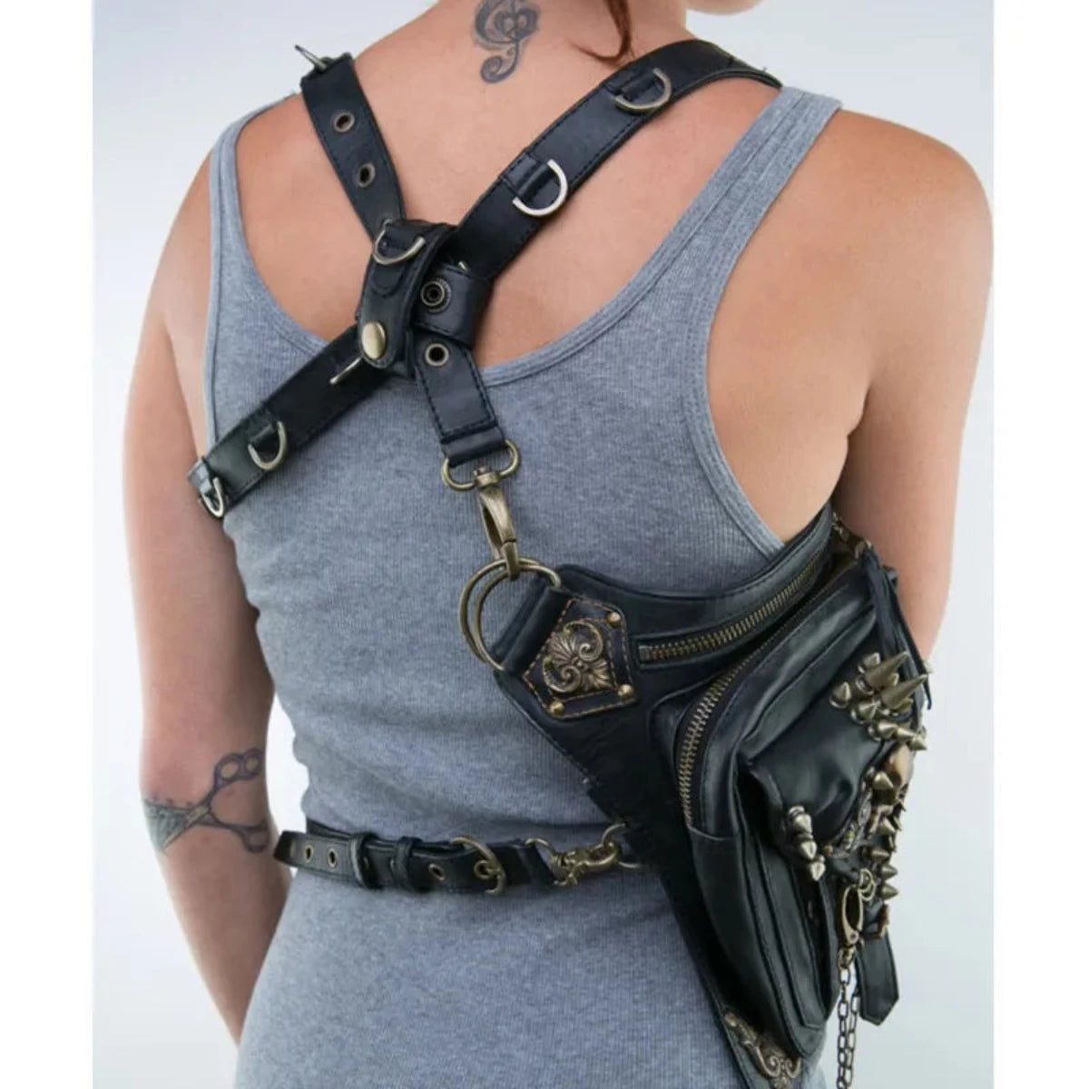 A person wearing a gray tank top is shown from the back, displaying a black leather harness with multiple straps, metal rings, and a zippered pouch. The Women's Gothic Waist/Hip/Body Fanny Bag is perfect for those who appreciate functional yet Gothic-inspired design. Tattoos are visible on their neck and arm.