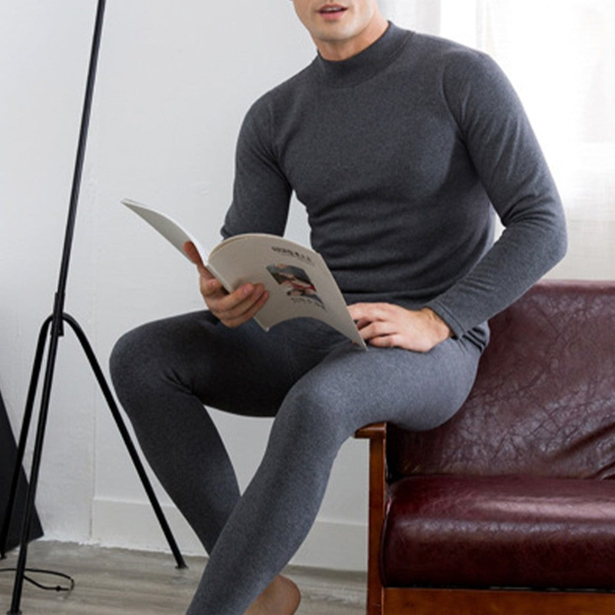 Sitting on a brown leather couch, the person is cozy in Men's Soft Winter Thermal Underwear - Dark Gray made from premium materials while holding and reading a magazine.