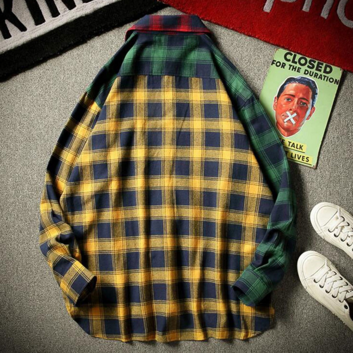 A Men's Finest Blended Color Flannel Shirt in red, with vibrant green and yellow checks, is showcased on a carpeted floor. Near it, a pair of white sneakers perfectly complements the outfit, while a colorful poster introduces an artistic touch. This standout piece from the men's clothing collection makes a stylish statement.