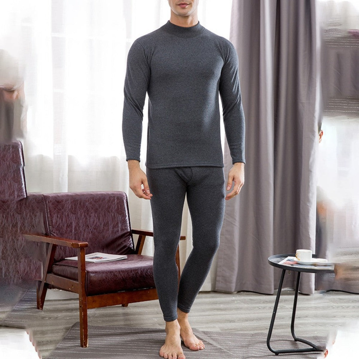 A person stands in a room, wearing Men's Soft Winter Thermal Underwear - Dark Gray. The premium long-sleeve top and matching bottoms provide warmth. A chair and small table with a cup and book are visible in the background.