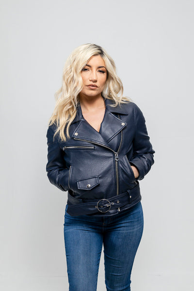 A woman wearing a First Manufacturing Remy - Women's Vegan Faux Leather Jacket, Navy Blue with an asymmetrical zipper and jeans standing against a plain background.