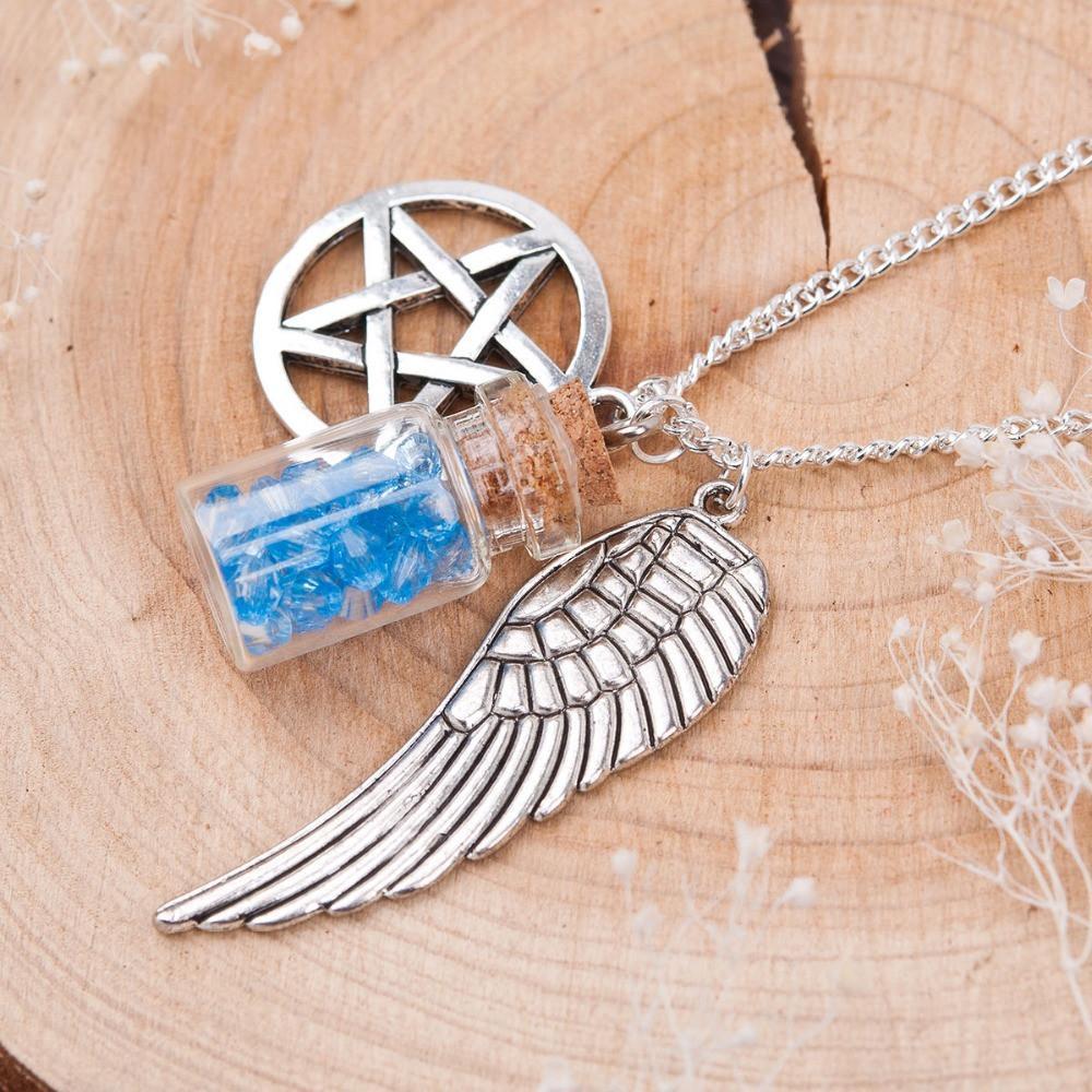 The Supernatural Pentacle Wings Necklace, showcasing a silver wing pendant, a gothic pentagram pendant, and a small bottle with blue crystals, rests elegantly on a wooden surface.