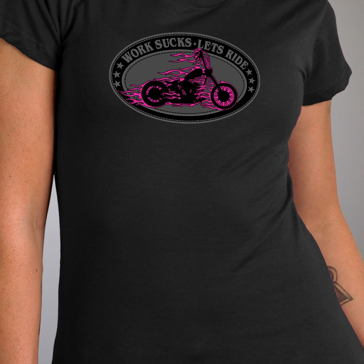 A person models the Hot Leathers Women's Work Sucks Oval Tee Shirt, a black t-shirt featuring metallic purple flames and a motorcycle graphic with the playful message "Work Sucks. Let's Ride" in pink—ideal for women who desire a garment that embodies boldness and adventure on every journey.