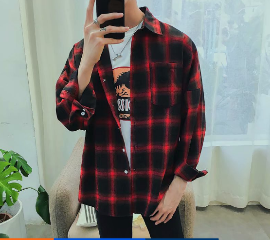 A person captures a mirror selfie wearing the Men's Classic Plaid Flannel Shirt in red and black, layered over a graphic tee. This stylish outfit is framed by the cozy setting of a room featuring a leafy plant and an inviting armchair.