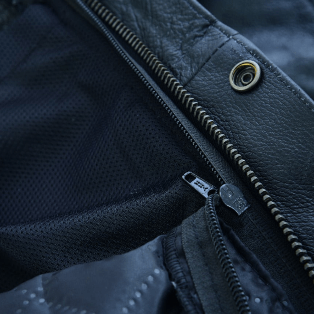 Close-up of the Vance Zip-Out Insulation Pant Style Motorcycle Leather Chaps, showcasing two zippers on black fabric and leather material reminiscent of classic chaps. One zipper is partially open, revealing a mesh lining beneath.