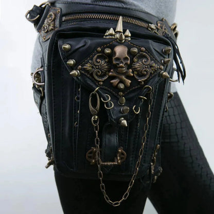 A Women's Gothic Waist/Hip/Body Fanny Bag in black leather, featuring ornate bronze skull and crossbones detail, chains, and metal studs. Worn on the side of a person sporting textured black leggings, this Gothic-inspired design offers spacious compartments for all your essentials.