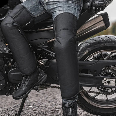 A person is seated on a motorcycle, dressed in black protective gear and jeans, highlighting the windproof design of the leg coverings that provide warmth similar to Alr™ Winter Motorcycle Riding Knee Warmers.