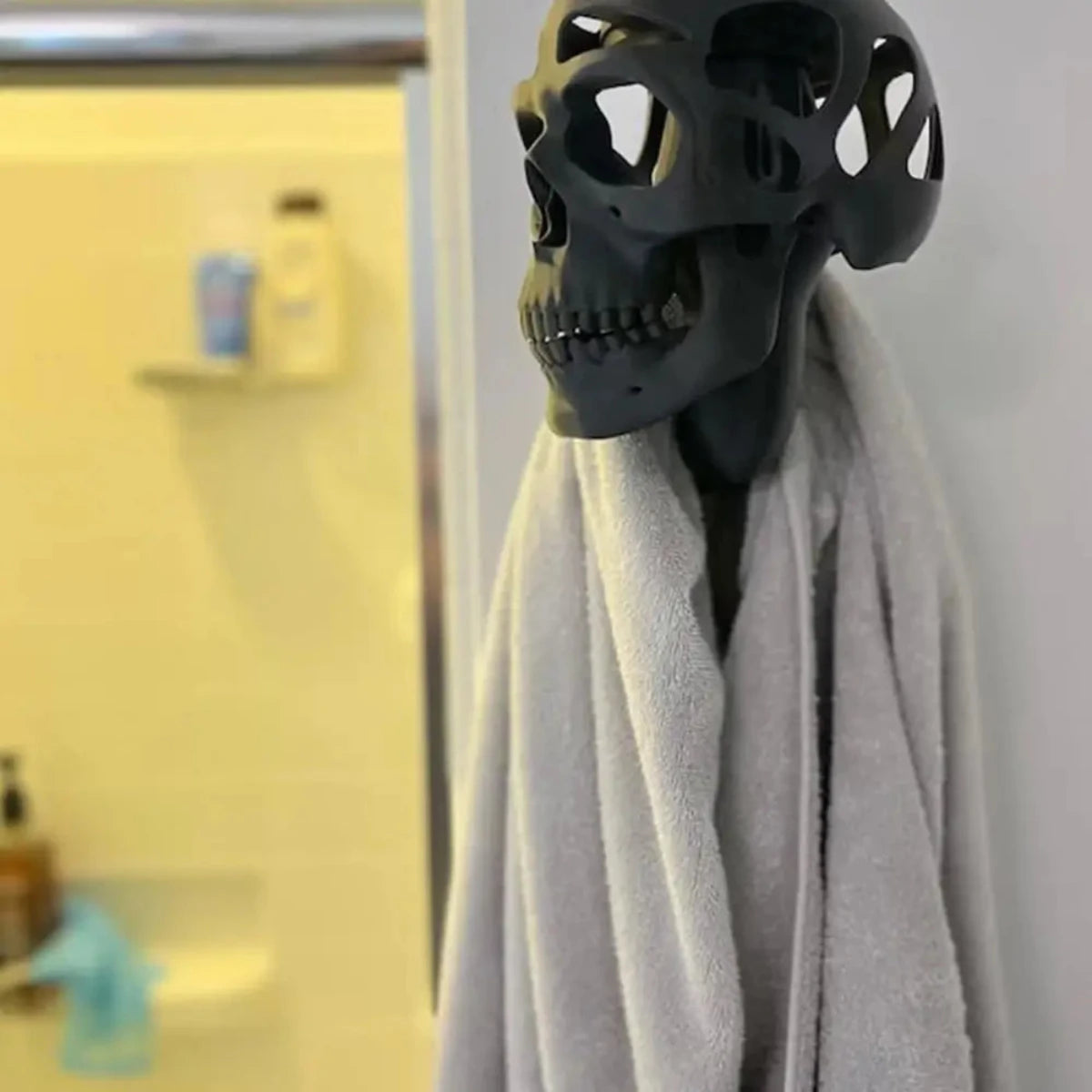 A gray towel hangs on an Alr™ 3D Skull Motorcycle Helmet Display Rack, shaped like a black skull, blending form and function in the bathroom with a shower visible in the background.