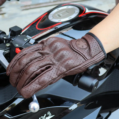 A gloved hand grips a motorcycle handlebar, showcasing the Alr™ Vintage Goatskin Full-Finger Motorcycle Glove in brown. This perforated leather accessory adds a classic touch while ensuring comfort. The motorcycle itself is black with red accents, enhancing the retro vibe.