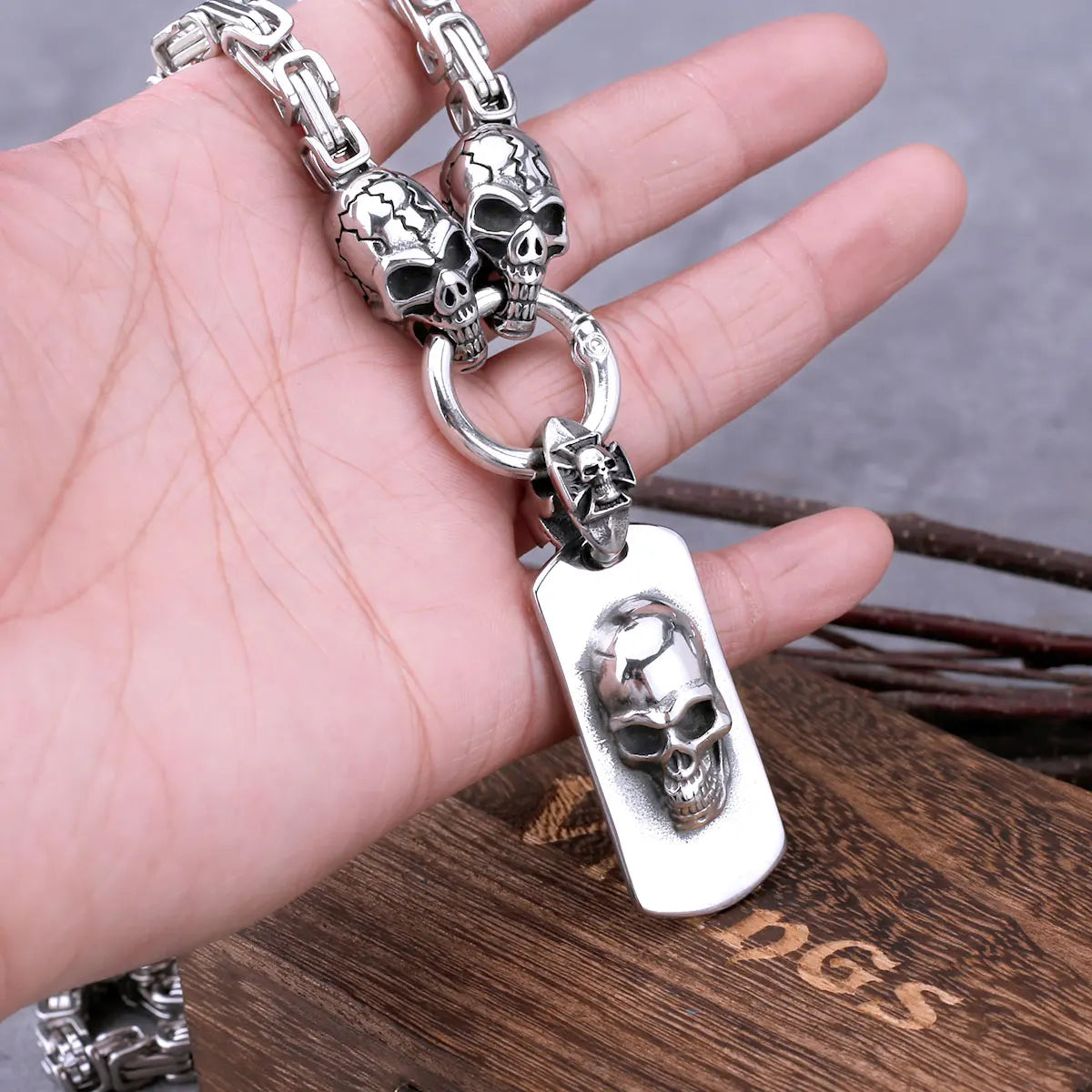 A hand holds the ALR™ Stainless Steel Pirate Skull Pendant Necklace, showcasing its silver design with a rectangular pendant and raised skull motif against a wooden box backdrop.