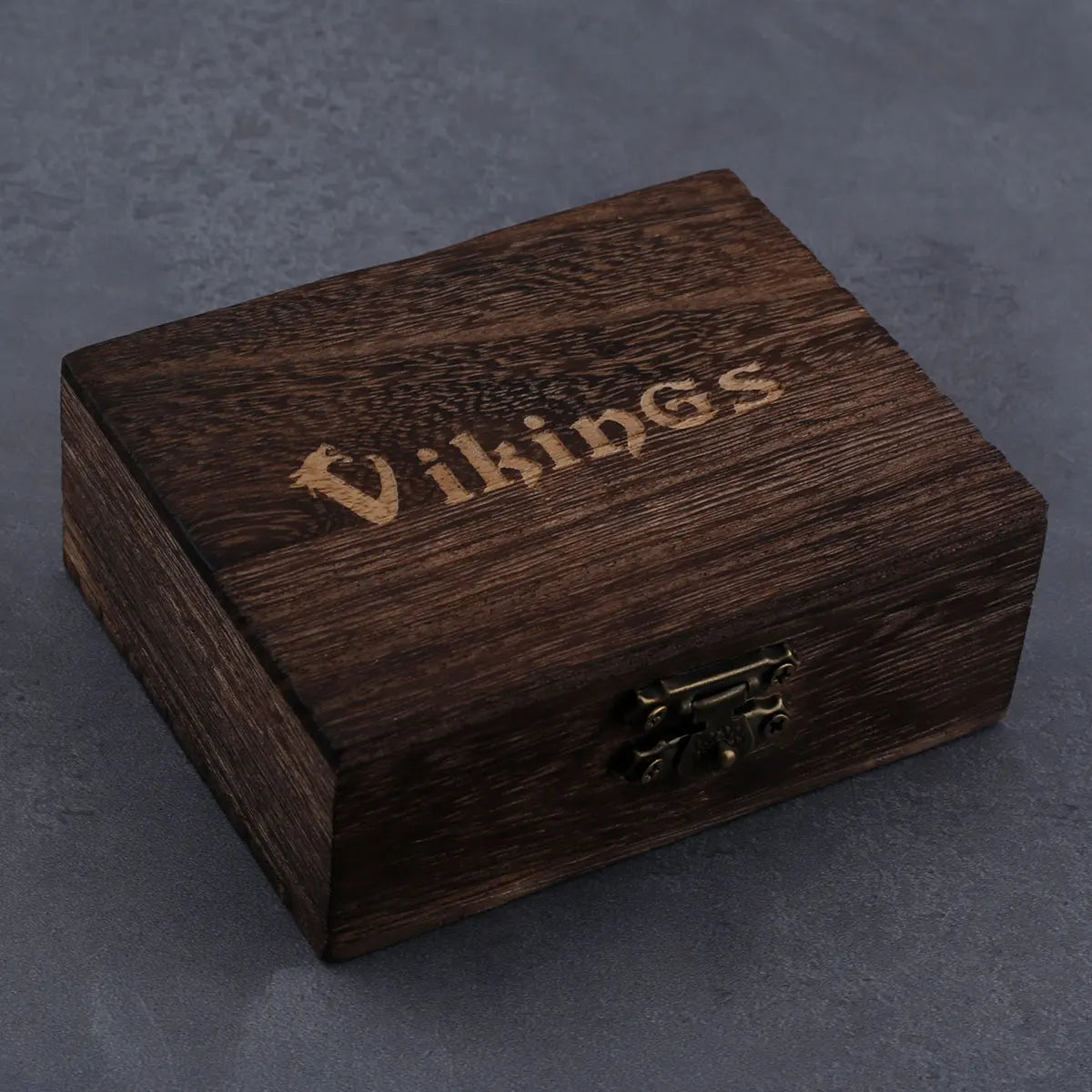 A closed wooden box, elegantly engraved with “Vikings” and secured with a metal latch, rests on a gray surface. Inside, you might discover vintage jewelry or an ALR™ Stainless Steel Pirate Skull Pendant Necklace to enhance your collection.