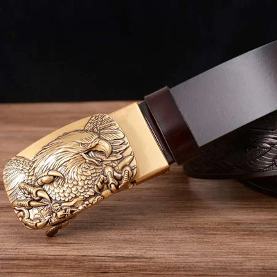 The Alr™ Men's Leather Ratchet Belt, crafted from genuine leather and featuring a gold automatic buckle with an engraved eagle design, rests elegantly on a wooden surface. Its versatile width makes it a stylish addition to any outfit.