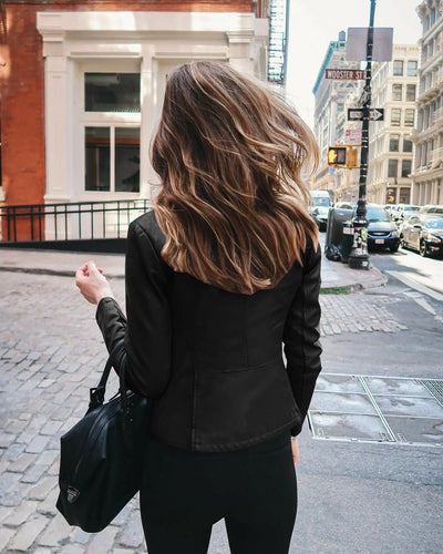 A woman with long hair exudes a moto biker vibe while striding down the city streets in an Alr™ Chic Lady Slim Fit Moto Jacket. She confidently carries a black bag against the backdrop of urban buildings and street signs.