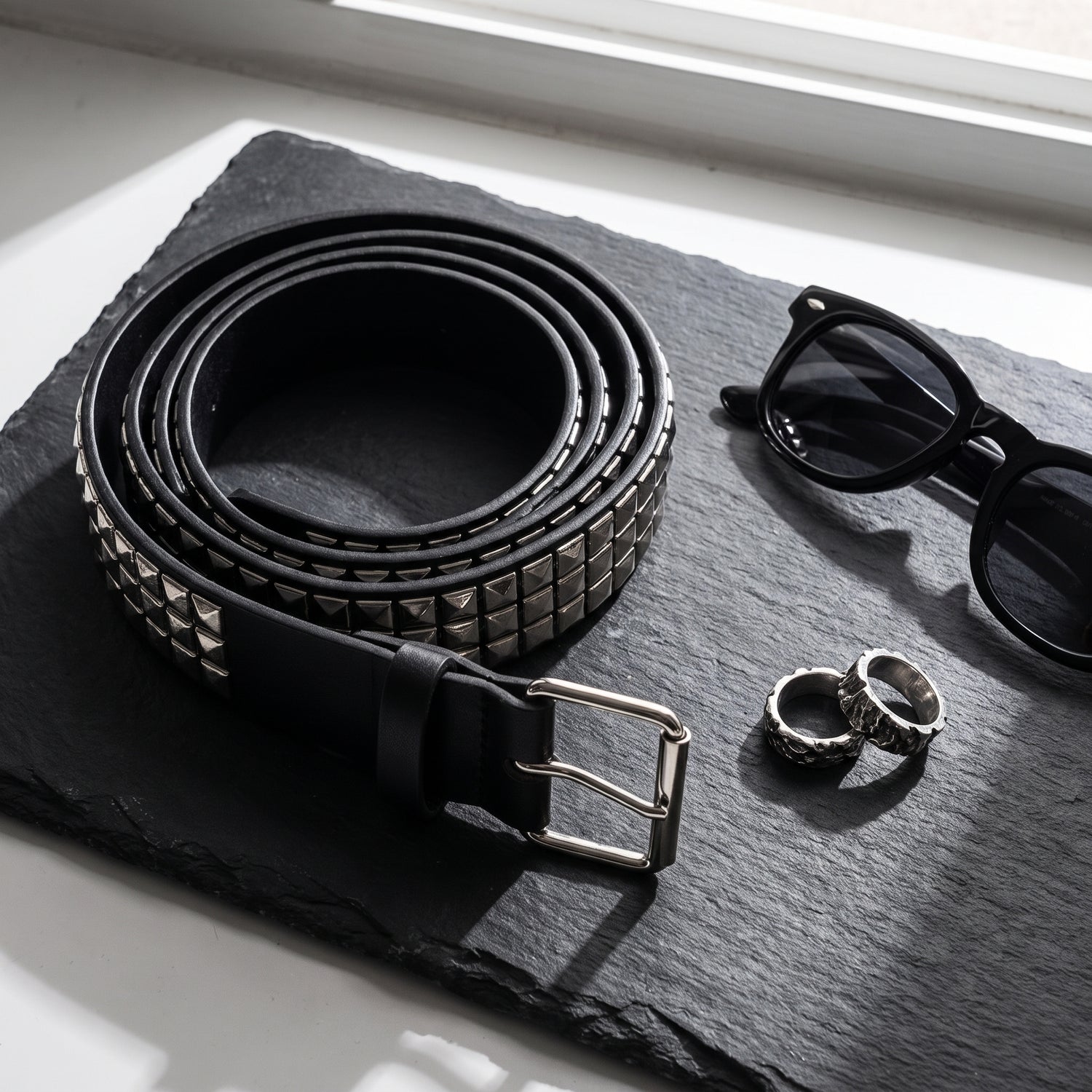Pyramid studded belt coiled on dark slate beside sunglasses and silver rings