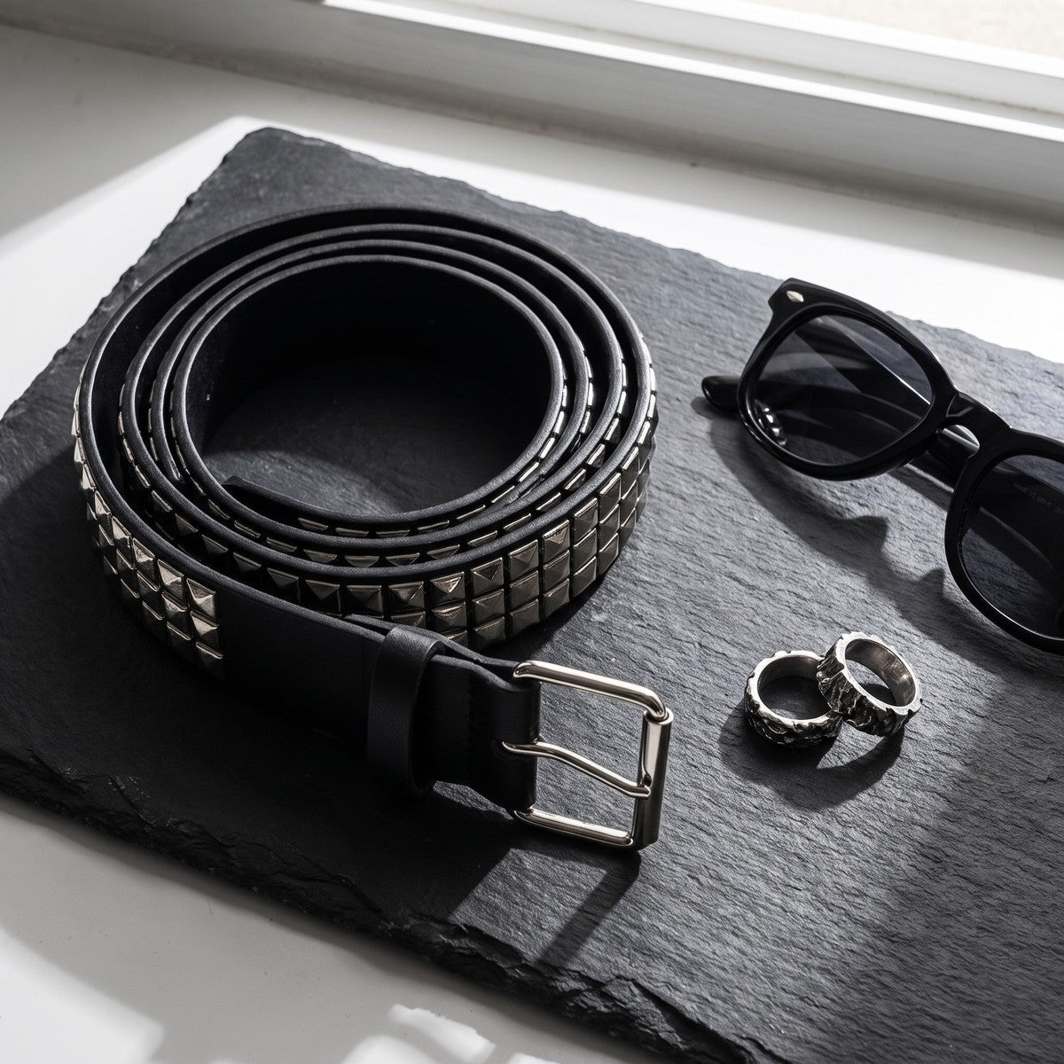 Pyramid studded belt coiled on dark slate with sunglasses and silver rings