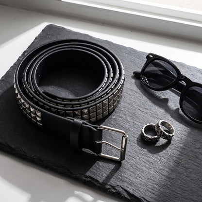 Pyramid studded belt coiled on dark slate with sunglasses and silver rings