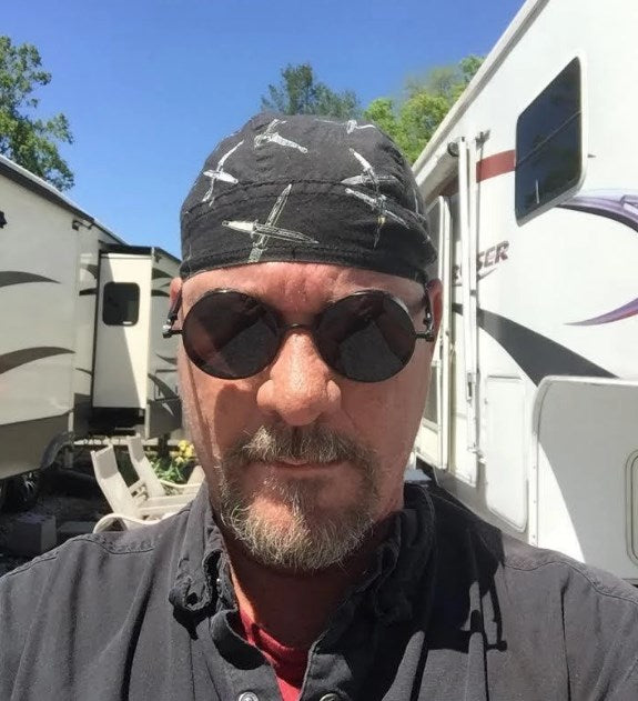 A biker wearing Rebel Steampunk Sunglasses plus Free 1% Er Ring Bundle and a bandana in front of an RV, these sunglasses not only serve as a fashion statement but also keep the wearer protected.