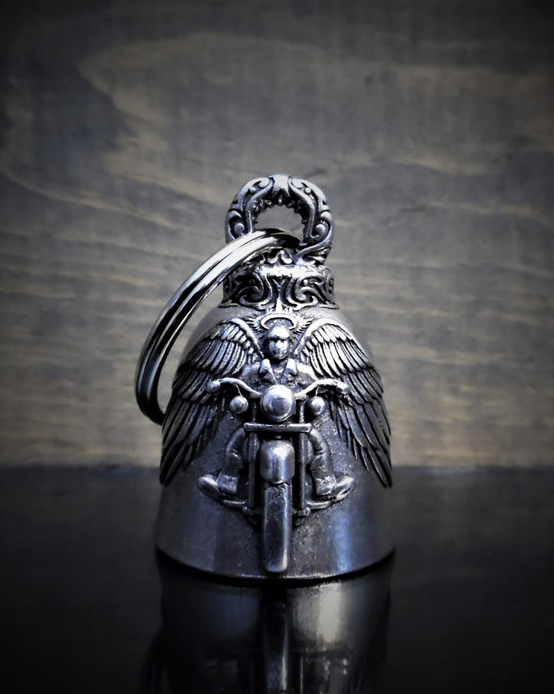 A small, intricately detailed bell with an engraved angel on a motorcycle, featuring wings spread wide. Crafted from lead-free pewter and branded as Daniel Smart Motorcycle Angel Bell, it comes with a keyring attached to the top. For added elegance, it's presented in a velveteen drawstring bag.