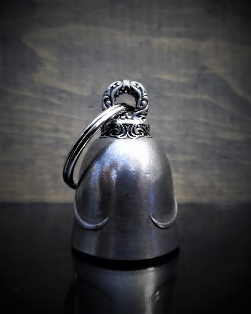 A small, metallic bell made of lead-free pewter with intricate carvings on its top loop, placed on a dark reflective surface against a blurred wooden background. This Daniel Smart Motorcycle Angel Bell comes nestled in a velveteen drawstring bag for added elegance.