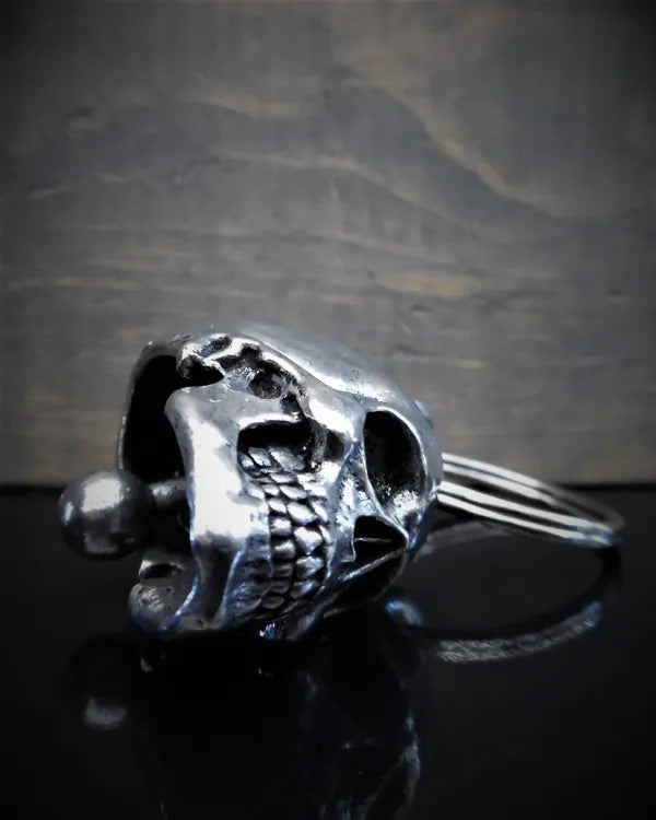 Close-up of a metal skull-shaped keychain crafted from lead-free pewter, lying on a glossy black surface with a blurred, dark background. The key ring is visible behind the skull. This Daniel Smart Skull Guardian Bell comes in a velveteen drawstring bag for added elegance.