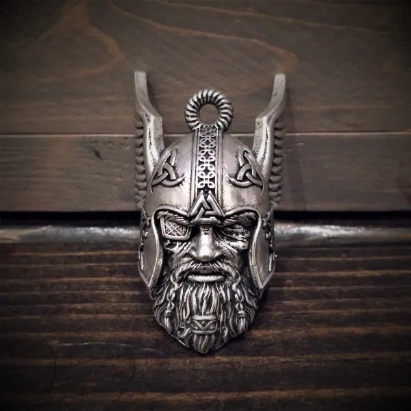 The Daniel Smart Odin Viking God Bell, a Viking warrior helmet pendant crafted from lead-free pewter with intricate carvings, is elegantly displayed against a wooden background and comes packaged in a velveteen drawstring bag by Bravo Bells.