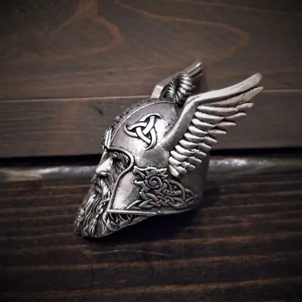 The Daniel Smart Odin Viking God Bell by Bravo Bells is a detailed, silver-toned pendant featuring a helmet with winged sides, ornate designs, and a bearded face. Crafted from lead-free pewter and set against a dark wooden background, it comes in an elegant velveteen drawstring bag.