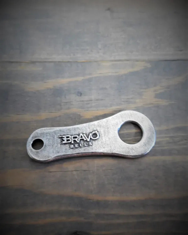 A small, flat metal tool made of lead-free pewter with the brand "BRAVO BELLS" engraved on it, featuring a hole on one end and a larger circular opening on the other, lies on a wooden surface. This USA-made item is popular among Daniel Smart Stars and Stripes Bell Hanger.