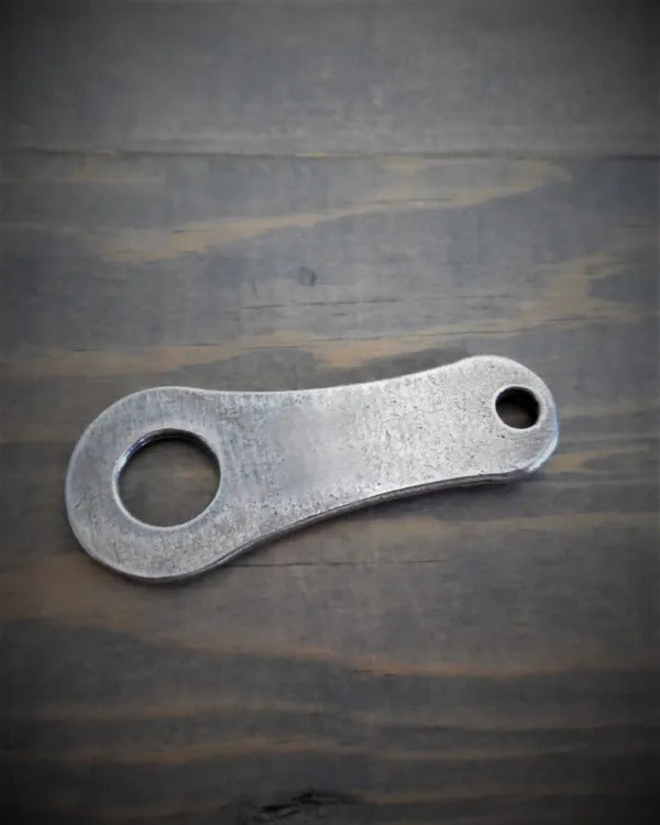 The Daniel Smart Bravo Bell Hanger is a lead-free pewter bottle opener with a simple, flat design featuring two different-sized circular holes on a wooden surface.