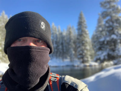 A person bundled in black winter gear, including a Daniel Smart Single Layer Fleece Neck Gaiter in black for multi-season use, stands outdoors with snowy trees and a river in the background.