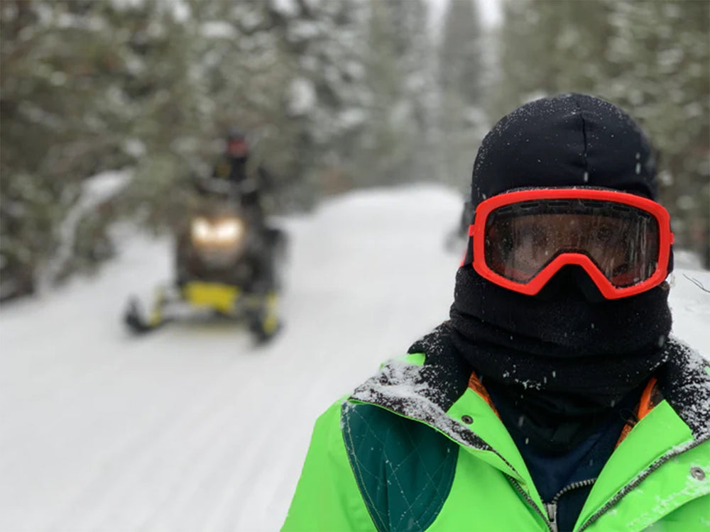 A person in a bright jacket and red goggles stands on a snowy path, warming up with a Daniel Smart Double Layer Fleece NeckGaiter in black. In the background, a snowmobile approaches amidst the trees.