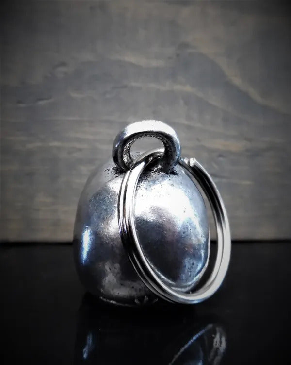 A small, metallic bell crafted from lead-free pewter with a smooth surface and a visible loop at the top, set against a dark background. This elegant piece, part of the Daniel Smart Skull Guardian Bell collection, comes nestled in a velveteen drawstring bag for safekeeping.