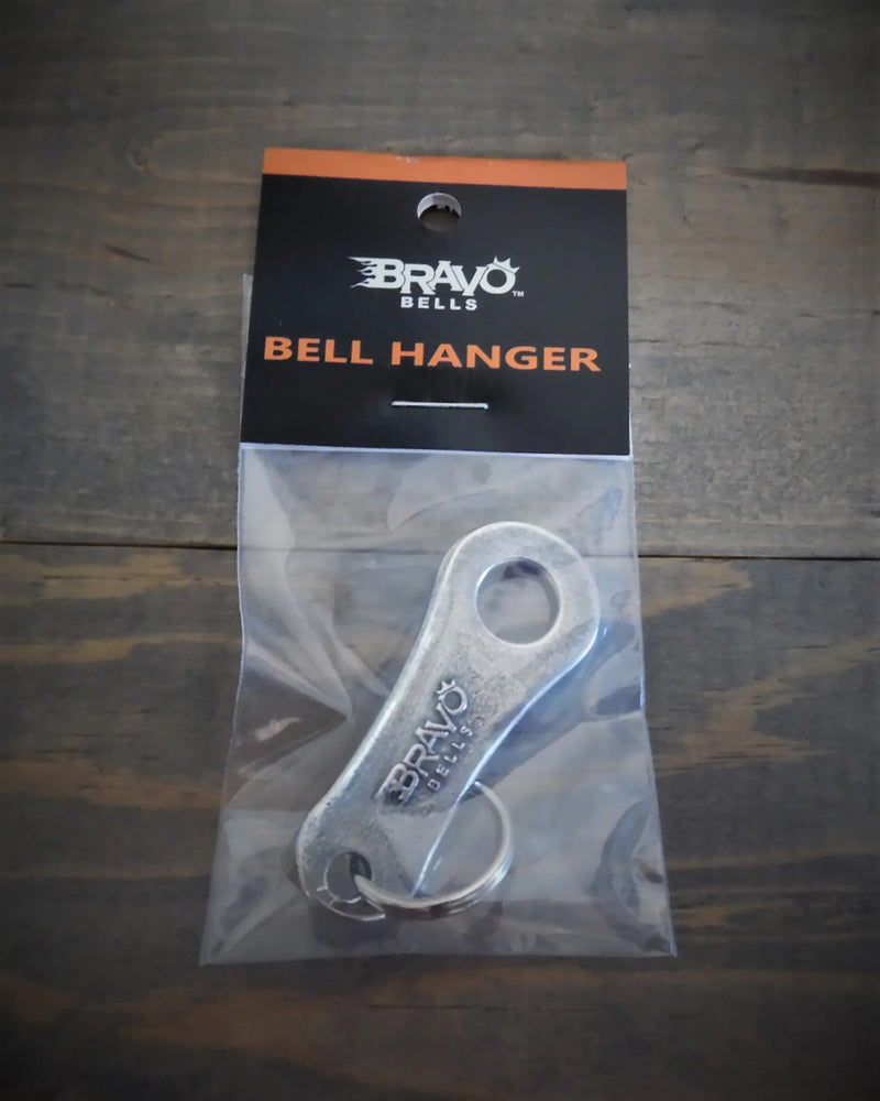 A packaged Daniel Smart Bravo Bell Hanger, crafted from lead-free pewter, is displayed on a wooden surface. The metallic tool features a sturdy split ring attached, ensuring durability and convenience.