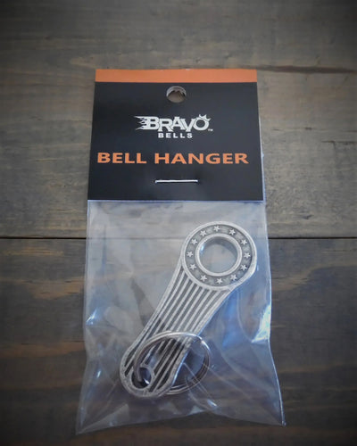 A Daniel Smart Stars and Stripes Bell Hanger in its packaging is placed on a wooden surface. The USA-made, lead-free pewter hanger is silver with star decorations and comes with a ring attachment.