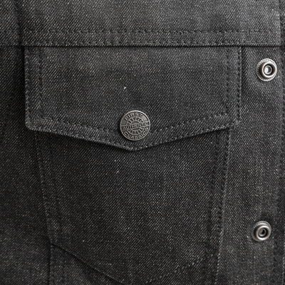 Close-up of a black denim vest pocket with a metal button. The button features an embossed design, and the vest, reminiscent of First Manufacturing Rook - Men's Motorcycle Denim Vest, showcases intricate stitching details.