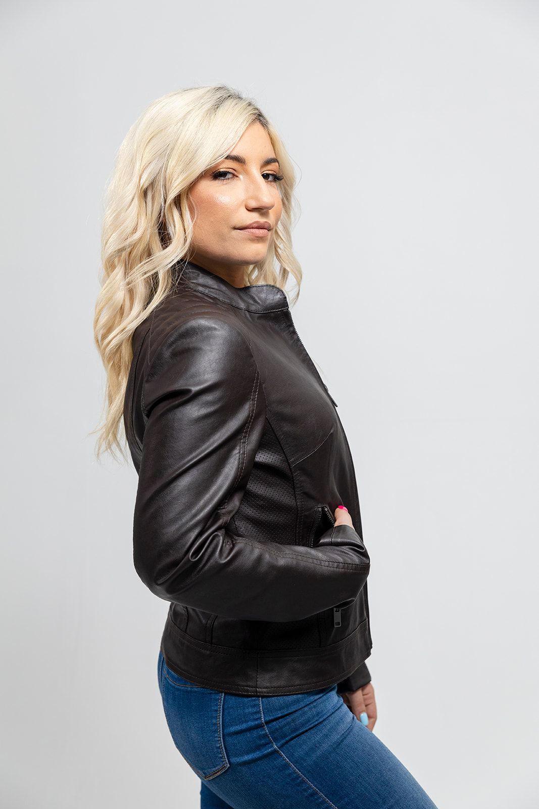 A woman is posing in jeans and the First Manufacturing Beverly - Women's Vegan Leather Jacket, Brown, showcasing fashion jacket style.