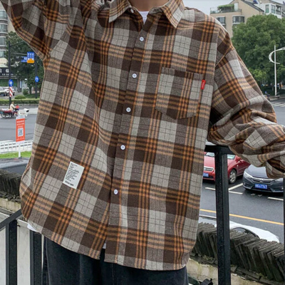 Person wearing an extra thick and cozy Men's Classic Plaid Flannel Shirt, Yellow in shades of brown and gray, with a red tag on the chest pocket and a white label on the lower front. Urban background with street and buildings.